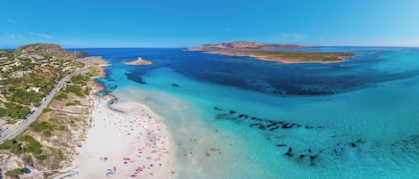 An aerial view of a coastal area with a beach, clear blue waters, and a road running parallel to the shore.