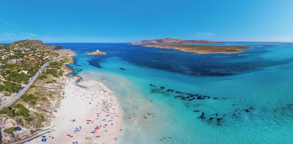An aerial view of a coastal area with a beach, clear blue waters, and a road running parallel to the shore.