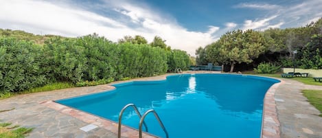 A rectangular swimming pool with a stone border, surrounded by a stone patio and lush greenery.