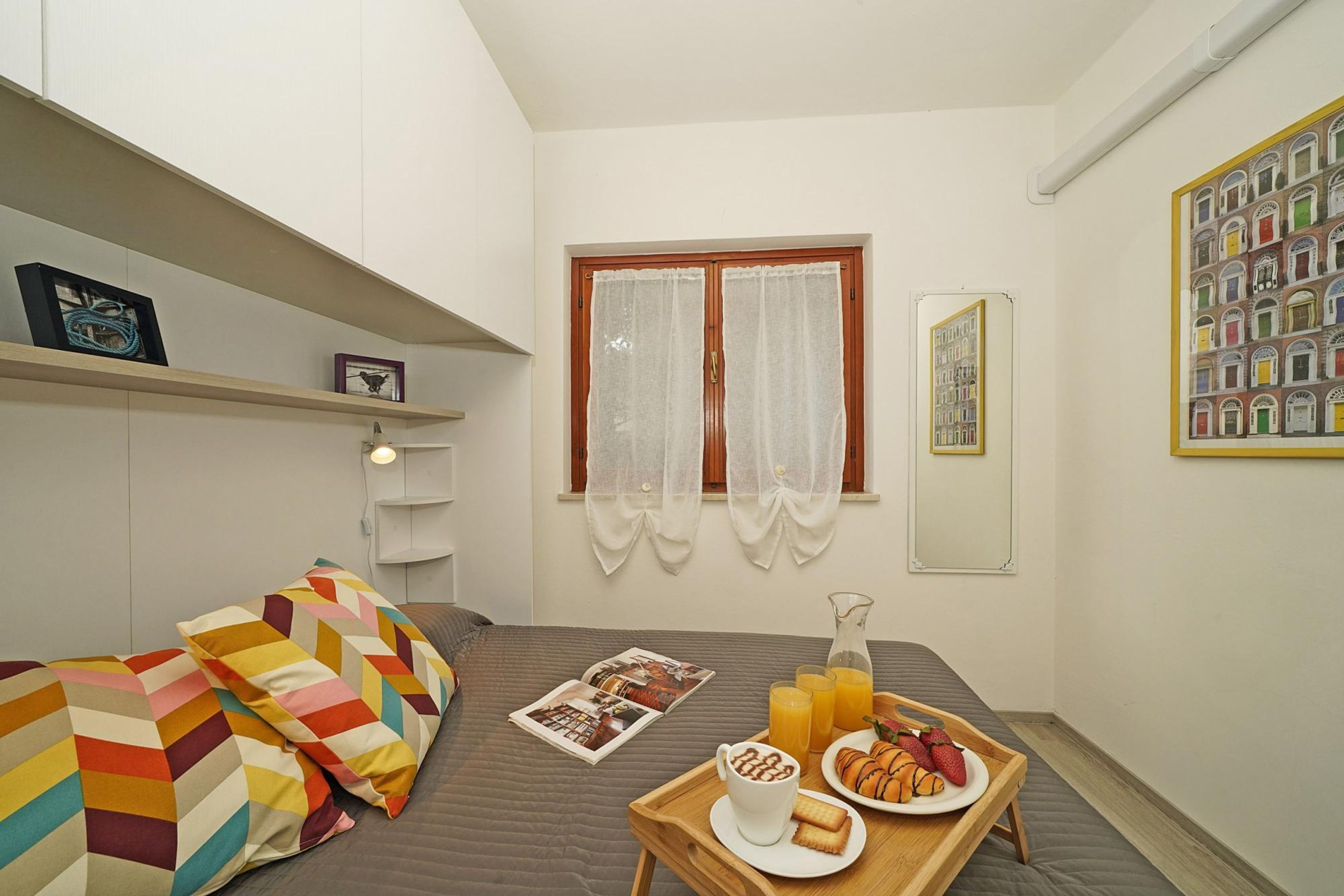 Une petite chambre à coucher, bien rangée, avec un lit, une table de chevet sur laquelle se trouve un plateau de petit-déjeuner, une fenêtre avec des rideaux et une étagère fixée au mur, ornée d’un tableau encadré.