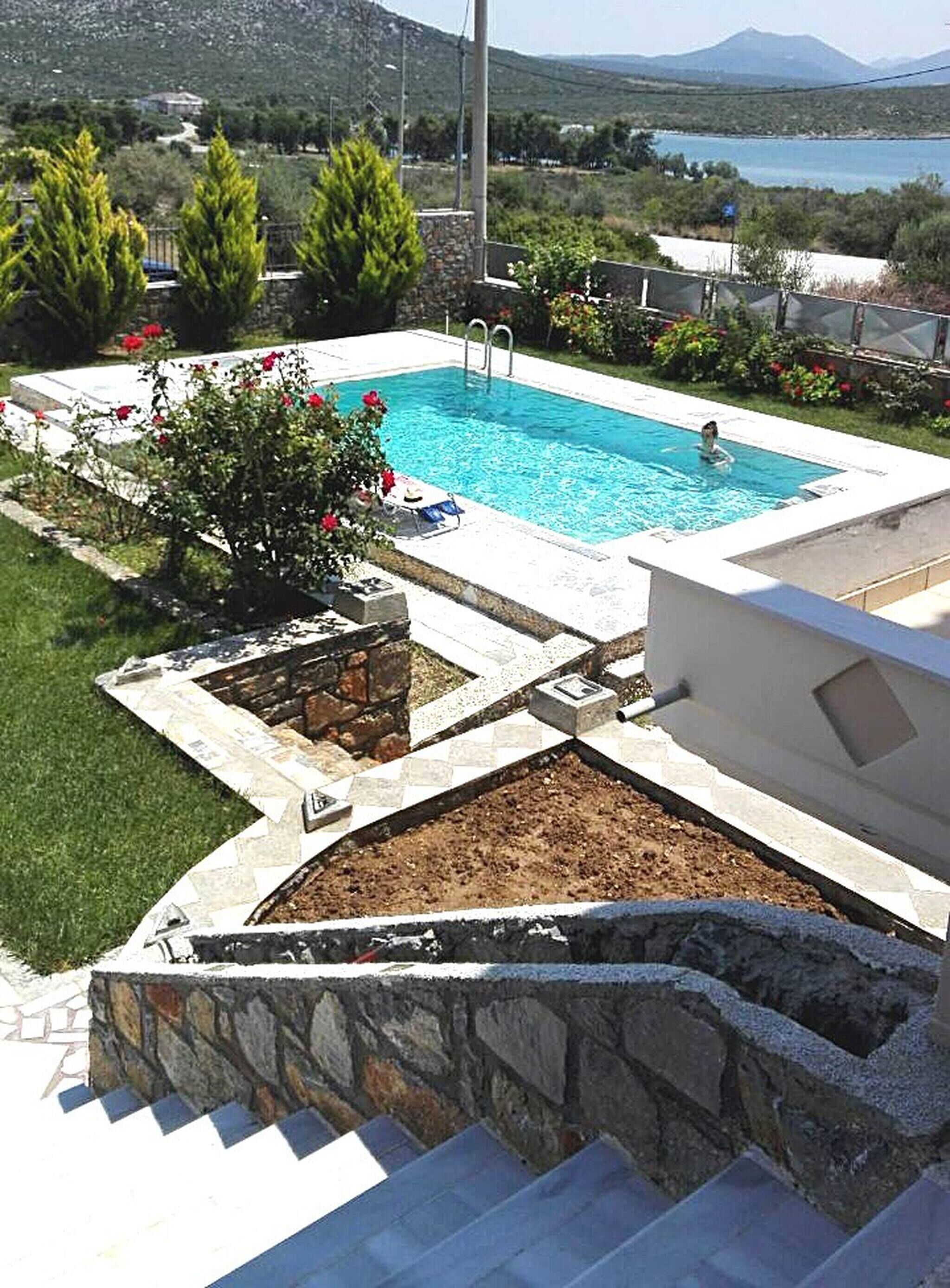 A rectangular swimming pool with a stone border, surrounded by greenery and a view of a lake and mountains.