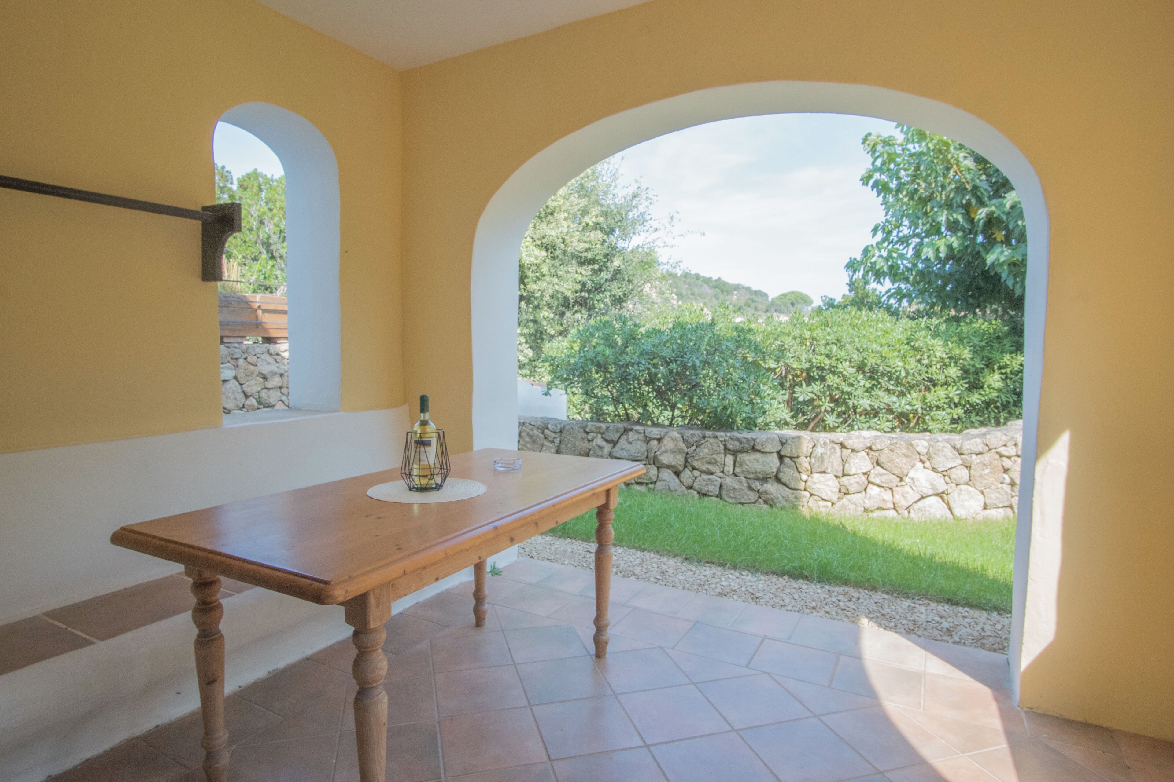 Ein Zimmer mit einem Holztisch, einer Flasche und einem Glas darauf. Zwei Rundbogenfenster bieten Blick auf Grünflächen und eine Steinmauer.