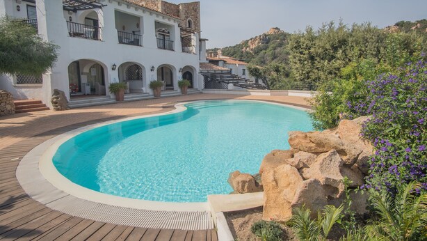 Una piscina con cubierta de madera, un edificio blanco con arcos y balcones, y montañas al fondo.