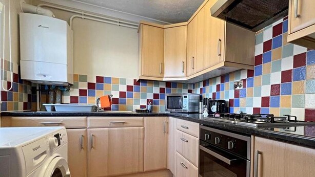 A compact kitchen with wooden cabinets, a washing machine, a microwave, a stove, and a colorful tiled backsplash. - 1 Bed Apartment, Sleeps 2, Free Parking, Tv (Enfield)