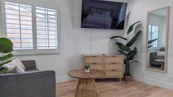 A living room with a flat-screen TV mounted on the wall, a round wooden coffee table, a grey sofa, a wooden cabinet, and large green plants.