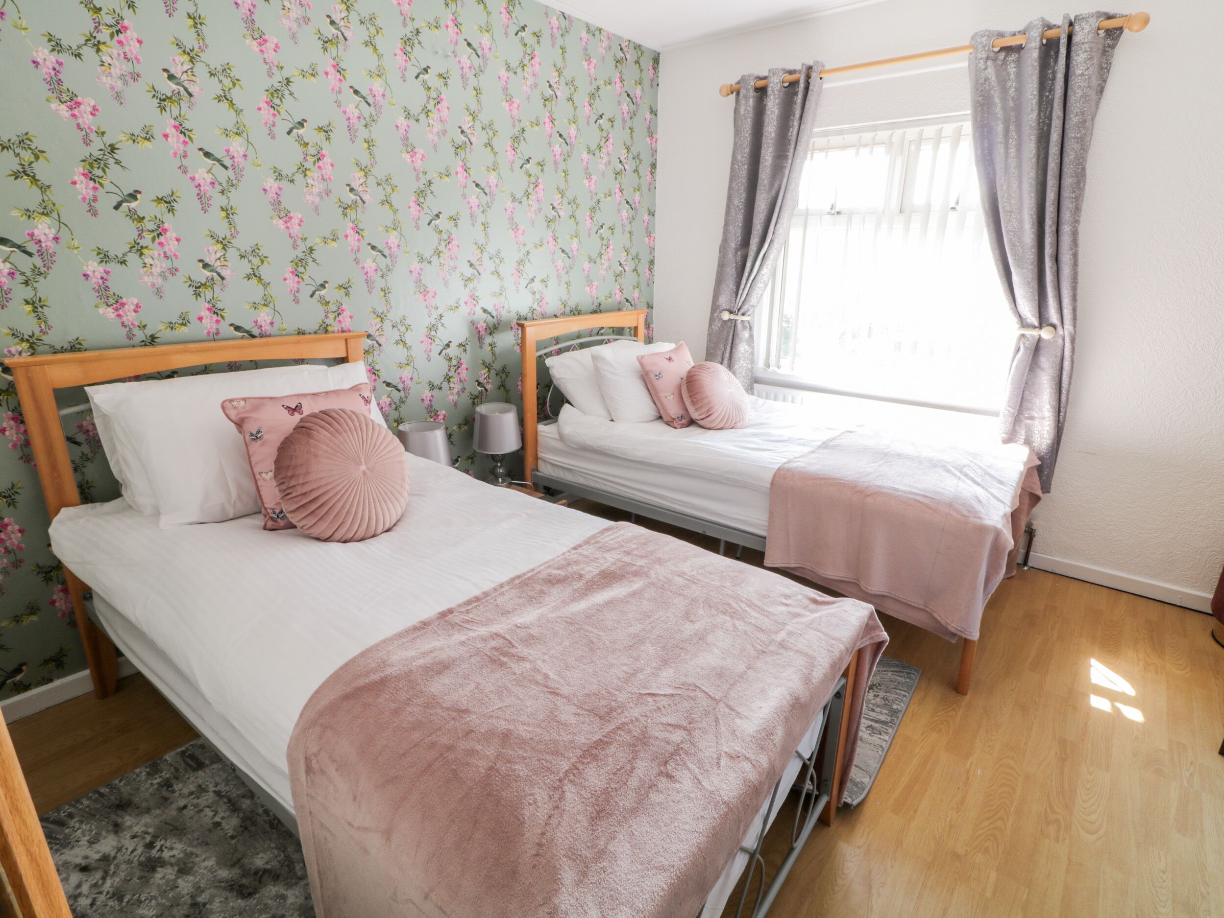 Een slaapkamer met twee eenpersoonsbedden, bloemenbehang, een houten hoofdbord, een grijze gordijn en een grijs vloerkleed.