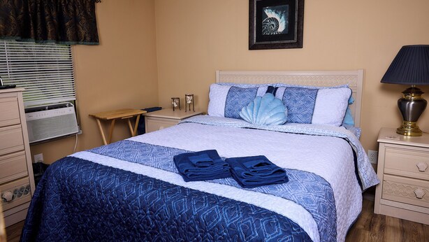 A neatly made bed with blue bedding, a bedside lamp, a framed picture on the wall, and a small table with a lamp.