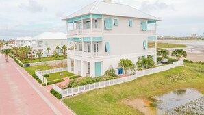 A multi-story coastal building with balconies, surrounded by a white fence and palm trees.