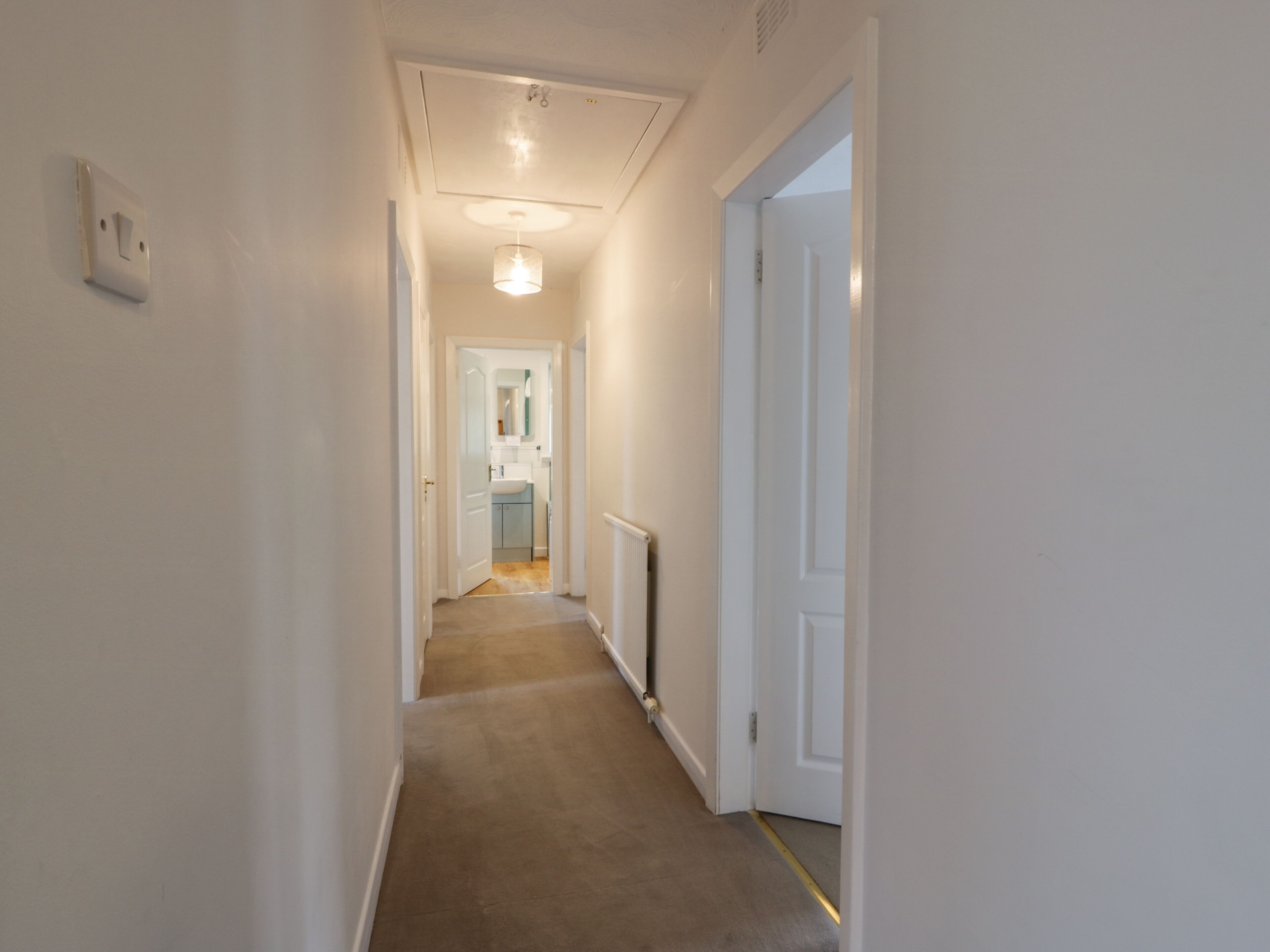 A narrow hallway with carpeted flooring, white walls, and a door leading to a bathroom with a sink and mirror.