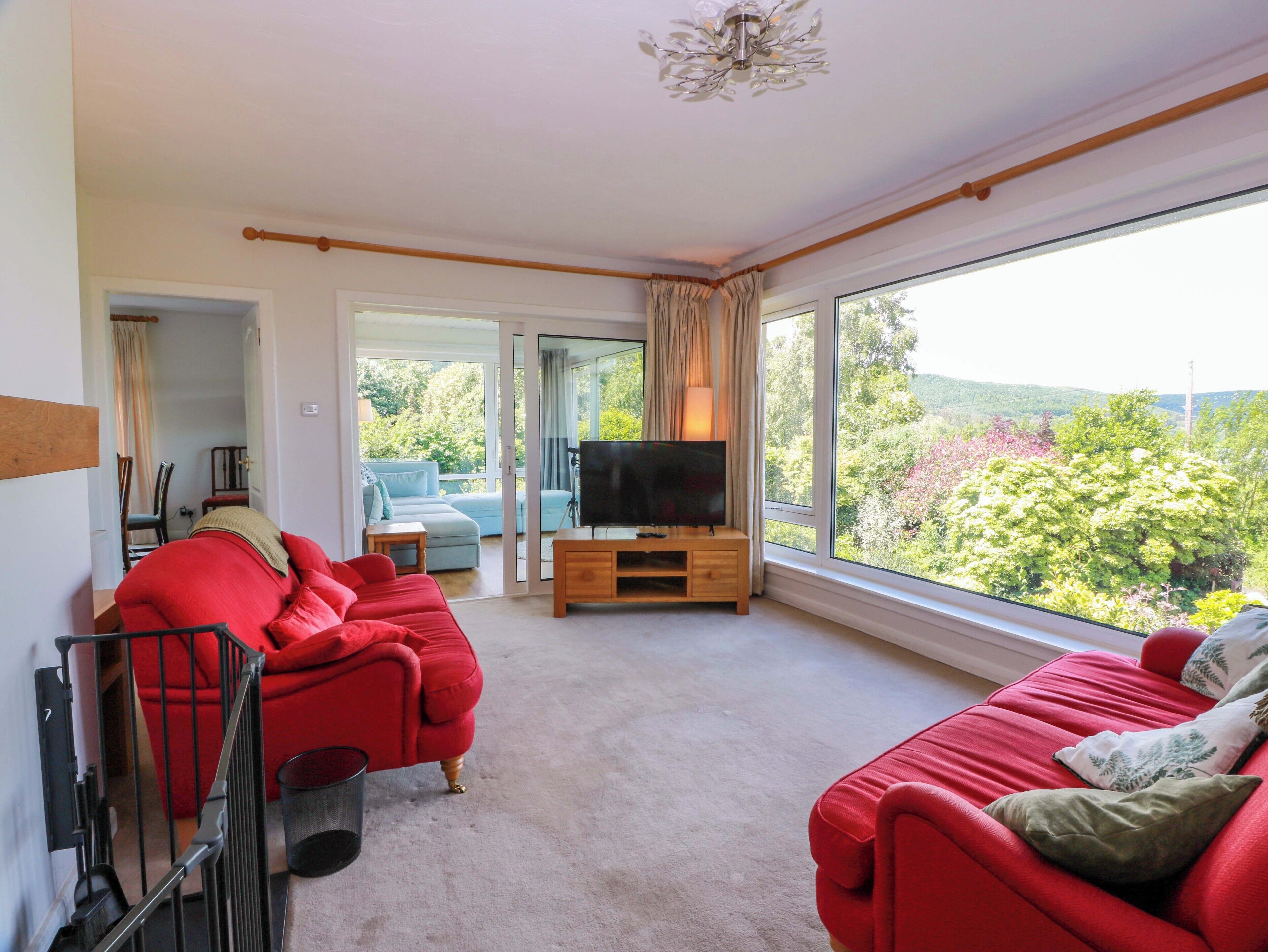 A spacious living room with a red sofa set, a television on a wooden stand, and a large window offering a view of greenery.