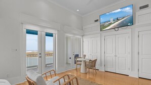 A spacious room with a large TV on the wall, a balcony with a view of the beach, and a dining area with wicker furniture. - Avalon - Townhouse At The Shores 4 Bedroom Home by RedAwning (South Padre Island)