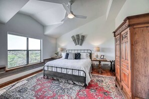 A bedroom with a large bed, a ceiling fan, a wooden wardrobe, a nightstand with a lamp, and a view through a large window. - Luxury 23-ac Ranch Hot Tub and Hill Country Views! (Fredericksburg)