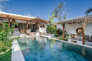 A modern outdoor pool area with lounge chairs, a thatched umbrella, and tropical plants. - Villa Ruta II (Tabanan)