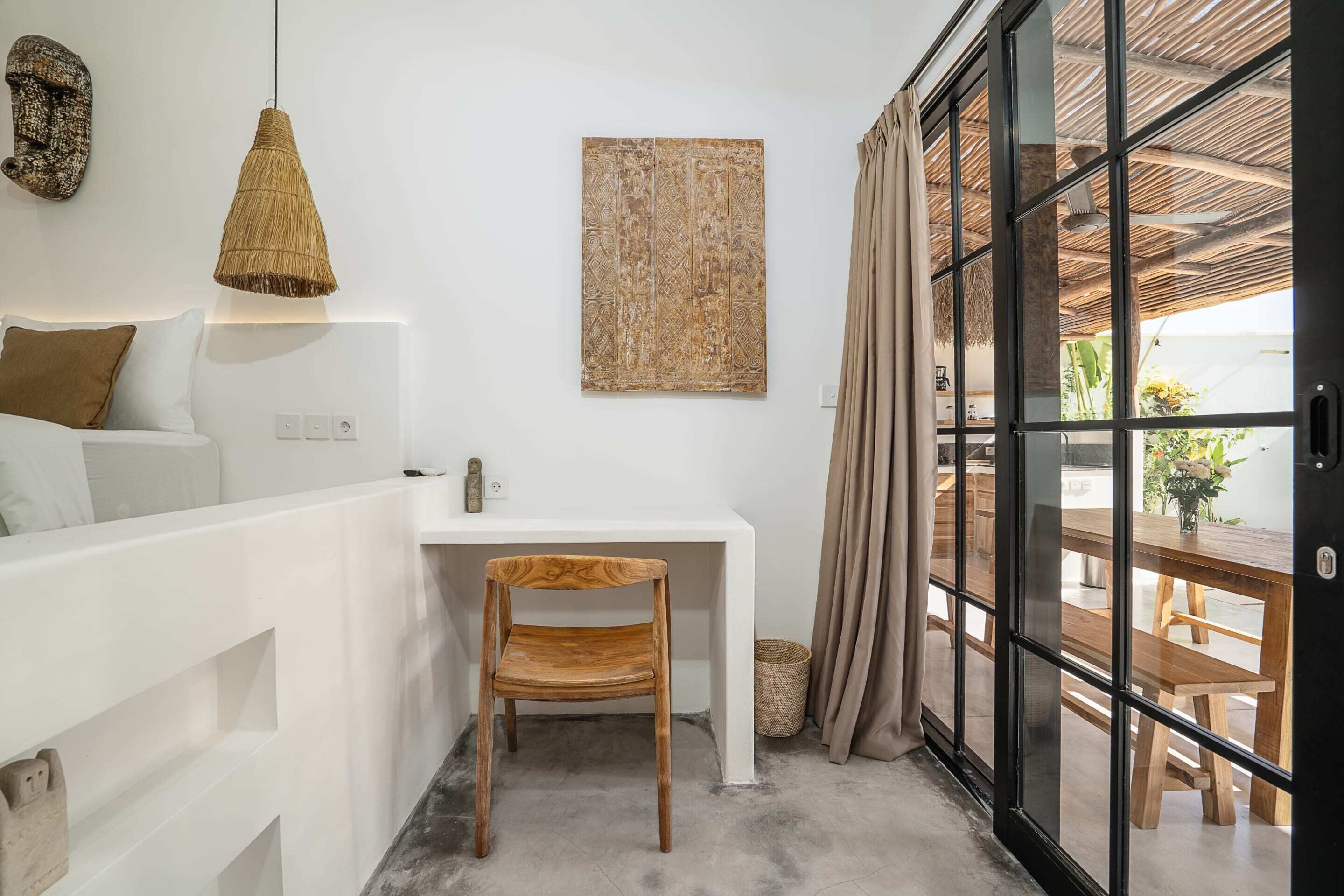 A room with a white desk, a wooden chair, a woven pendant light, a textured wall art piece, and a sliding glass door leading to an outdoor area with wooden furniture.