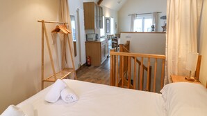A bedroom with a bed, a wooden ladder, and a hanging rack. There is a kitchen area visible through an open door. - The Loft Cottage (Totnes)