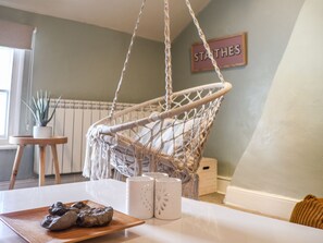 Une pièce avec un fauteuil suspendu en macramé, une petite table sur laquelle se trouve un plateau en bois ainsi que des salières et des poivrières, et une plante en pot.