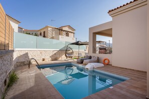 A swimming pool with a stone wall, a lounge area with a sofa and chairs, and a view of neighboring buildings.