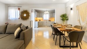 A modern living room with a sofa, dining table, and chairs. There is a kitchen in the background with cabinets and a hanging light fixture. - Villa Ismini 3 Bedrooms With Pool , sea View and E-car Charger (Rethymno)