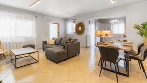 A modern living room with a sectional sofa, a dining table with chairs, and a kitchen area in the background. - Villa Ismini 3 Bedrooms With Pool , sea View and E-car Charger (Rethymno)