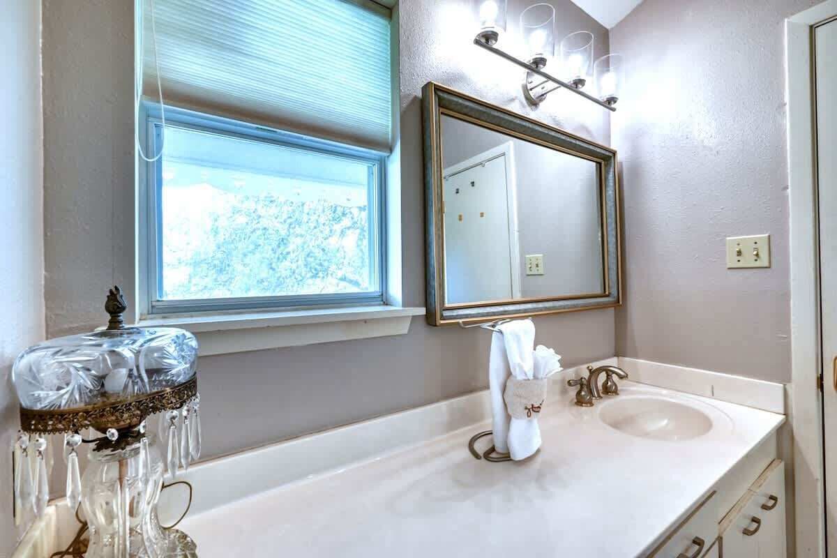 A bathroom with a large mirror, a window with blinds, a sink with a towel rack, and a decorative glass vase.