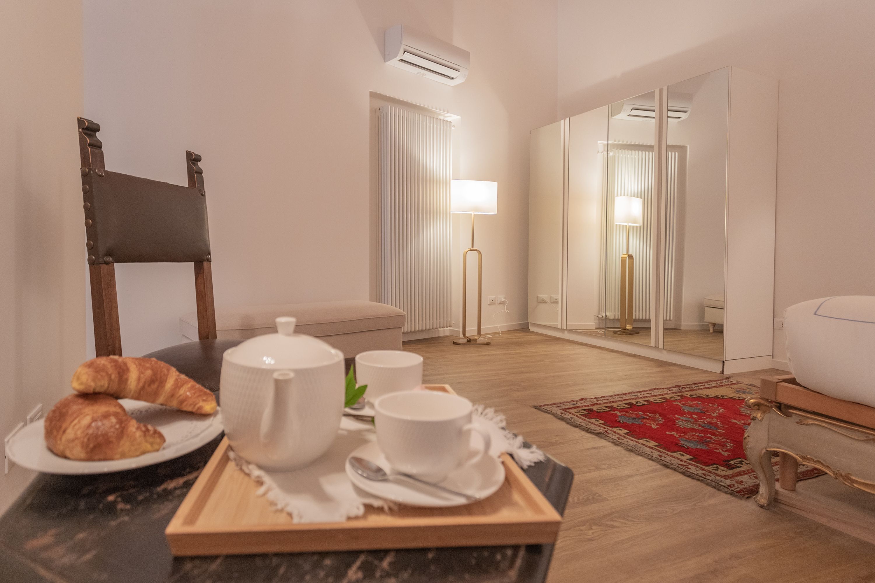 A room with a dining table set with croissants, a teapot, cups, and a tray. There is a chair, a floor lamp, and a mirror.