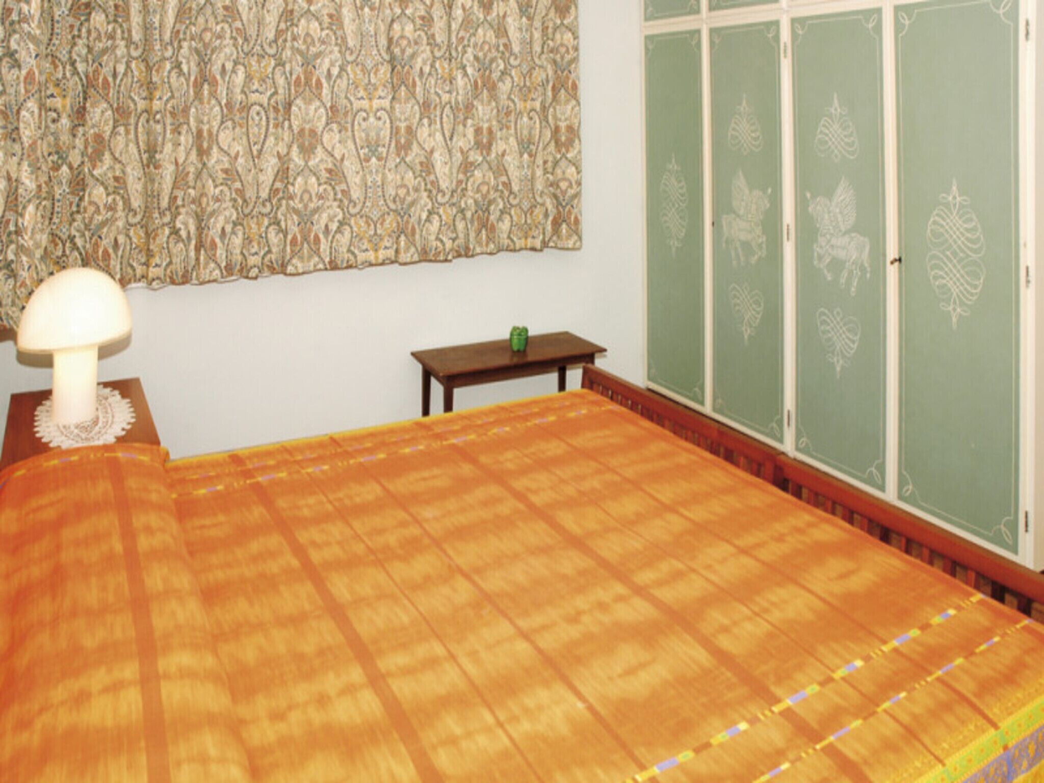 A room with a bed, a small table, a lamp, and green doors with decorative patterns.