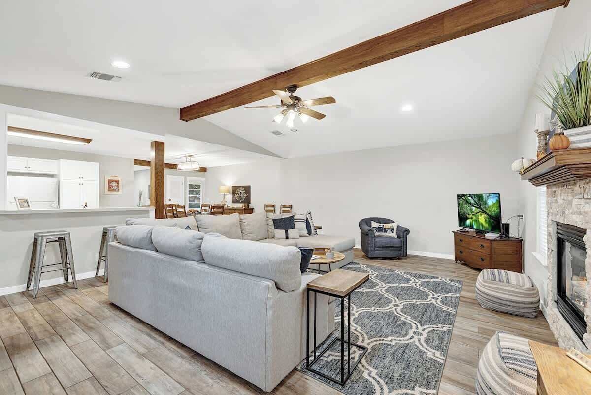 A spacious living room with a large gray sofa, a coffee table, and a fireplace. There is a television and a stone fireplace mantel.