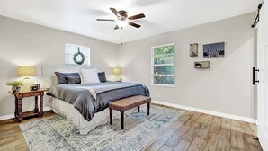A bedroom with a bed, two bedside tables, a ceiling fan, a window with blinds, and a door. - The Billie Goat Haus - Hot Tub -steps to Main St! (Fredericksburg)