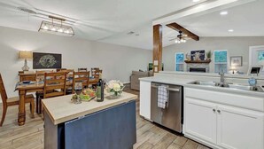 A modern kitchen with white cabinetry, a wooden dining table, and a built-in island with a sink. - The Billie Goat Haus - Hot Tub -steps to Main St! (Fredericksburg)