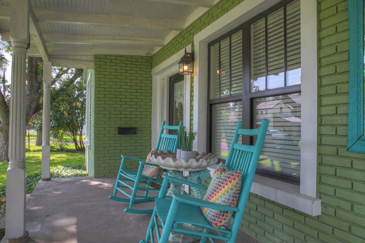 A porch with teal rocking chairs and a small table, green brick walls, and a window with shutters.