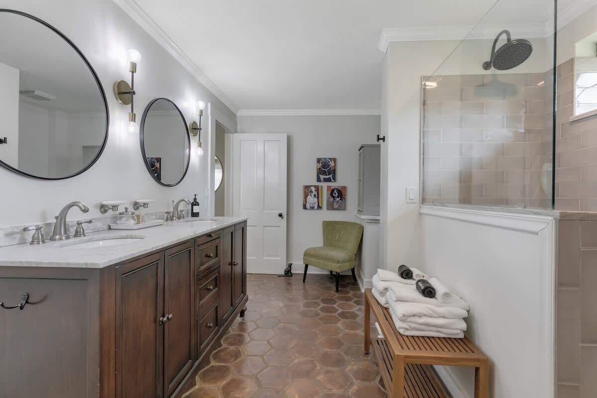 A bathroom with a large mirror, two sinks, a shower, and a chair.