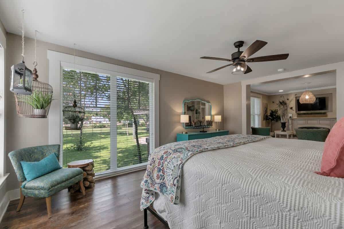 A bedroom with a large bed, a ceiling fan, a view of the outdoors through a sliding door, and a small seating area with a chair and a table.