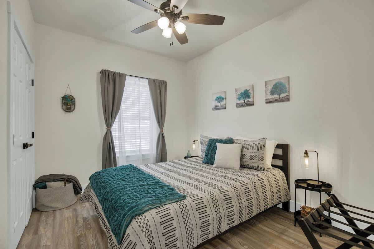 A bedroom with a bed, a ceiling fan, a window with curtains, a bedside table, and a wall-mounted lamp.