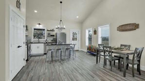 A spacious kitchen with a central island, bar stools, and a dining area with a wooden table and chairs. - Hill Country Hideaway With Fire Pit and Hot Tub! (Fredericksburg)