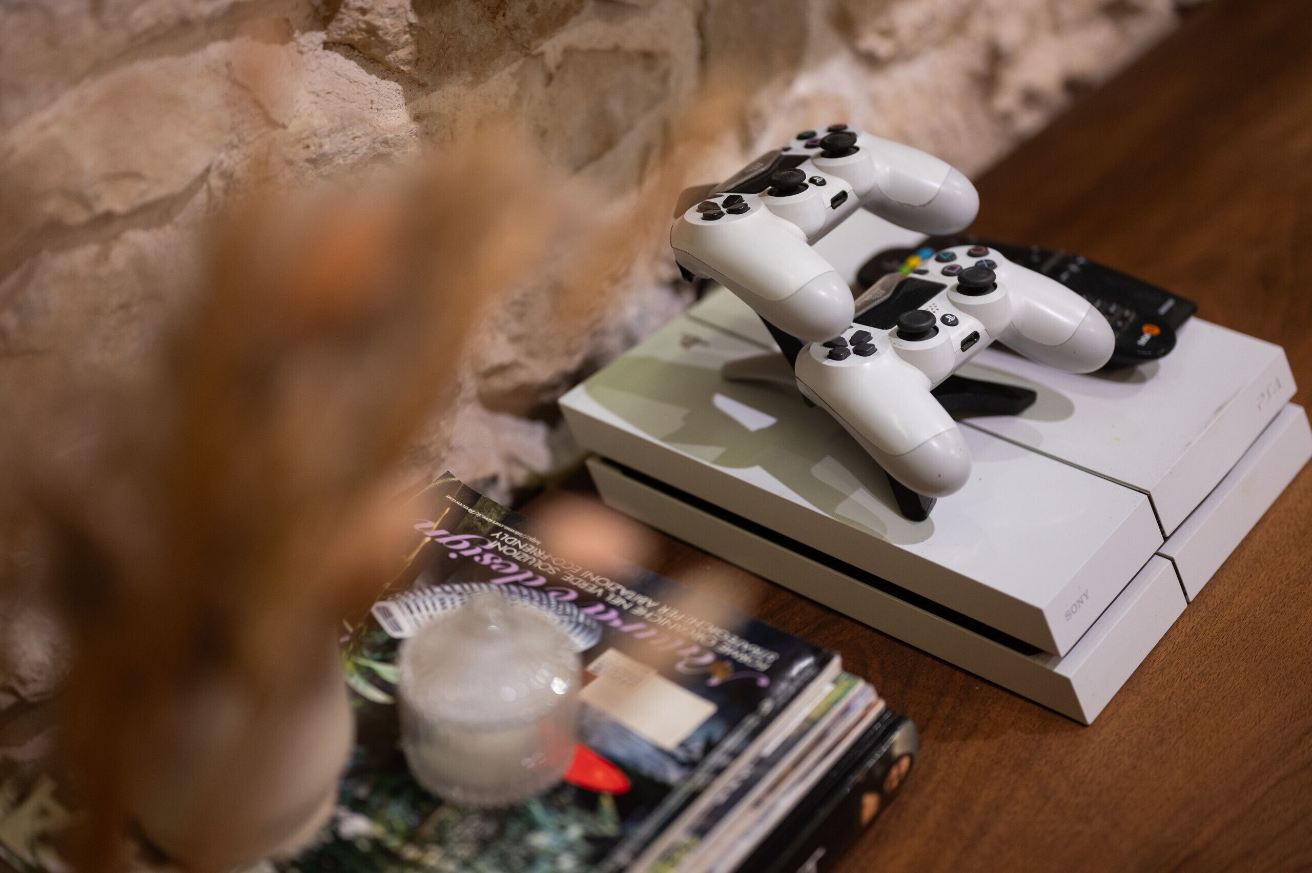 A white PlayStation 5 console with controllers on a wooden surface, surrounded by magazines and a small figurine.