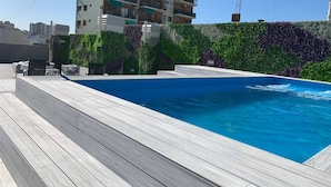 A swimming pool with a wooden deck, surrounded by a green wall with purple flowers, and a multi-story building in the background. - Brand New Apartment In Caballito (Buenos Aires)