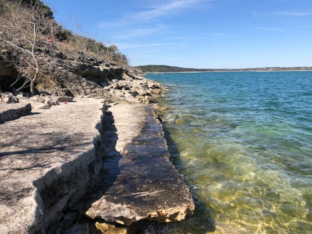 A rocky shoreline with clear water, a concrete barrier, and a distant hill.
