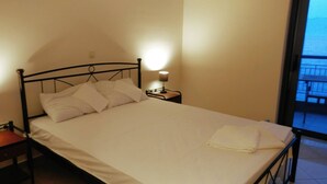 Una cama con ropa blanca, una mesita de noche con lámpara y vistas a un balcón a través de una ventana.