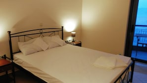 A bed with white bedding, a nightstand with a lamp, and a view of a balcony through a window. - The Pearl Seaview Apartment, 10sec Walk To The Sea (Messini)