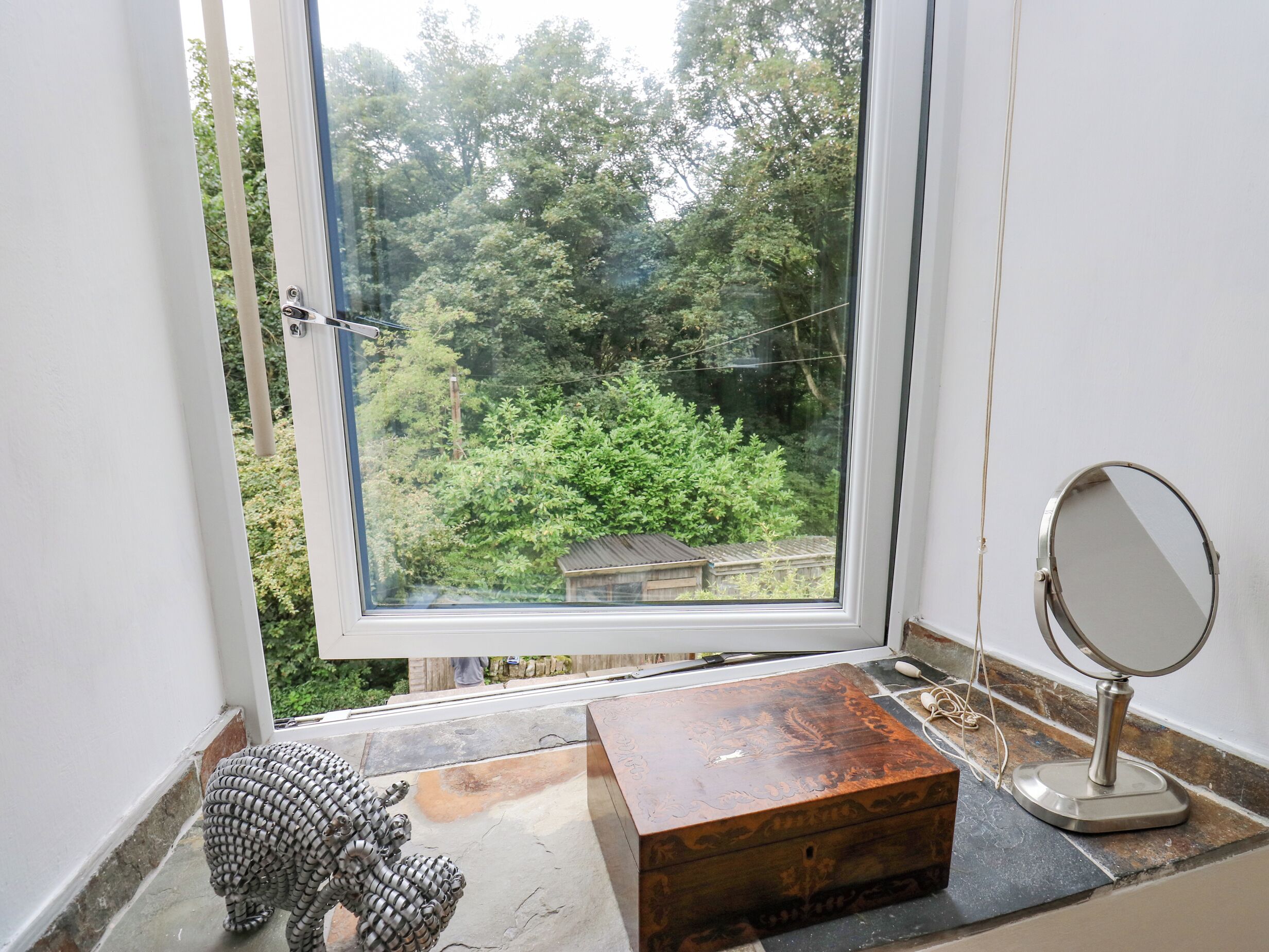 A room with a large window, a wooden chest, a small statue, and a round mirror on a tiled floor.