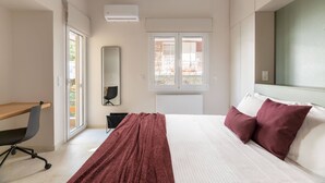 A bedroom with a bed, a desk, a chair, a mirror, and a window with blinds.