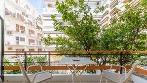 A balcony with two white metal chairs and a small table, overlooking a residential area with white buildings and green trees.