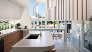 Une cuisine moderne dotée d’un grand îlot central, d’un coin repas avec des sièges extérieurs, et offrant une vue sur la piscine et le jardin.