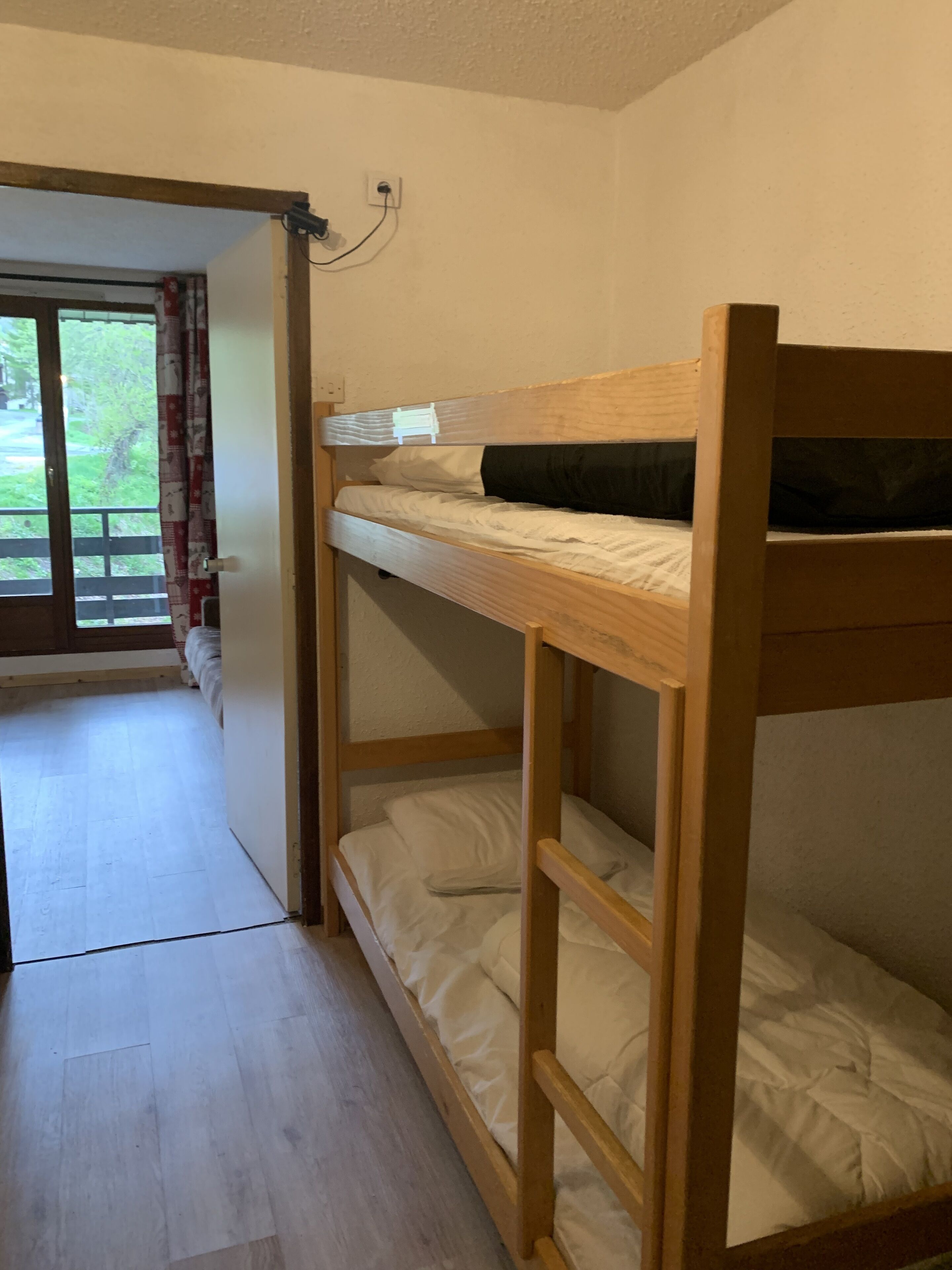 Een slaapkamer met stapelbed met houten frame, wit beddengoed en een deur naar een balkon met uitzicht op groen.