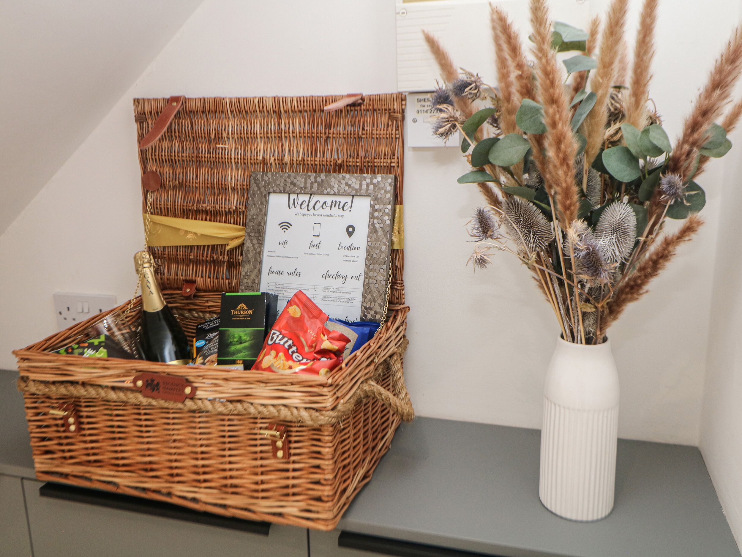 A wicker basket with a welcome sign, a bottle of champagne, and snacks on a counter next to a vase with dried plants.