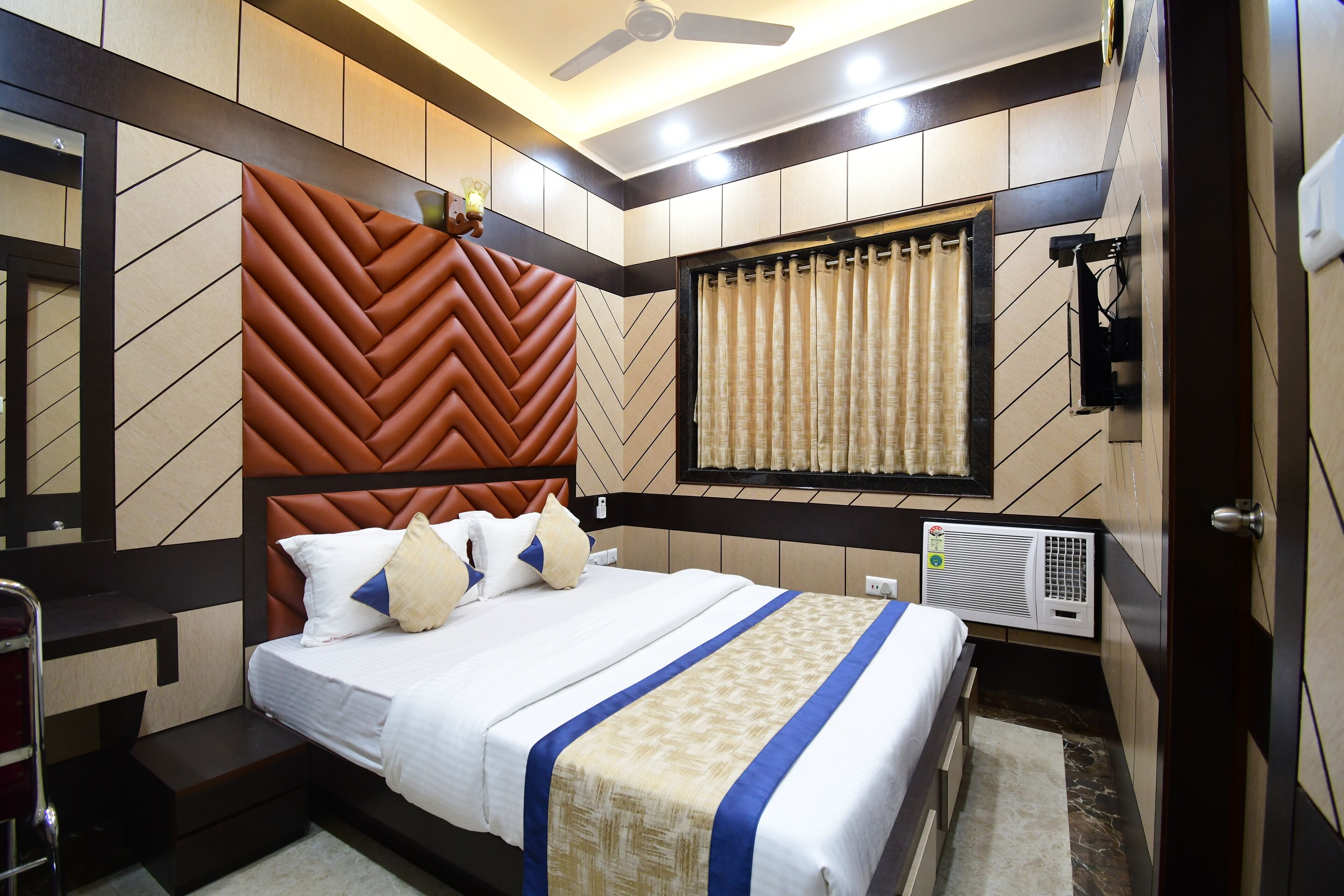 Una habitación de hotel moderna con una cama grande, cabecera con estampado en zig-zag, ventana con cortinas, equipo de aire acondicionado y ventilador de techo.