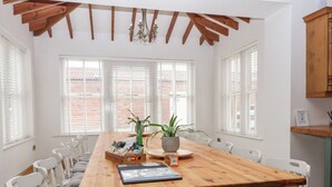 Un espace repas comprenant une table en bois, des chaises blanches, un lustre et de grandes fenêtres équipées de stores.