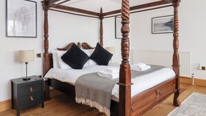 Un lit à baldaquin avec une structure en bois foncé, du linge de lit blanc et deux oreillers noirs. Il y a une petite table de chevet avec une lampe et un tiroir. La pièce est dotée d’un parquet et d’un radiateur blanc.