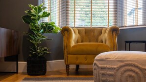 Una habitación con un sillón amarillo, un reposapiés blanco, una planta en maceta y una ventana con persianas.
