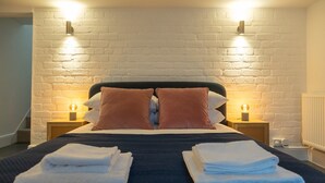 Una cama bien hecha con un cabecero oscuro, dos almohadas y toallas dobladas sobre un edredón azul oscuro. Hay dos mesitas de noche con lámparas y una pared de ladrillo blanco detrás de la cama.